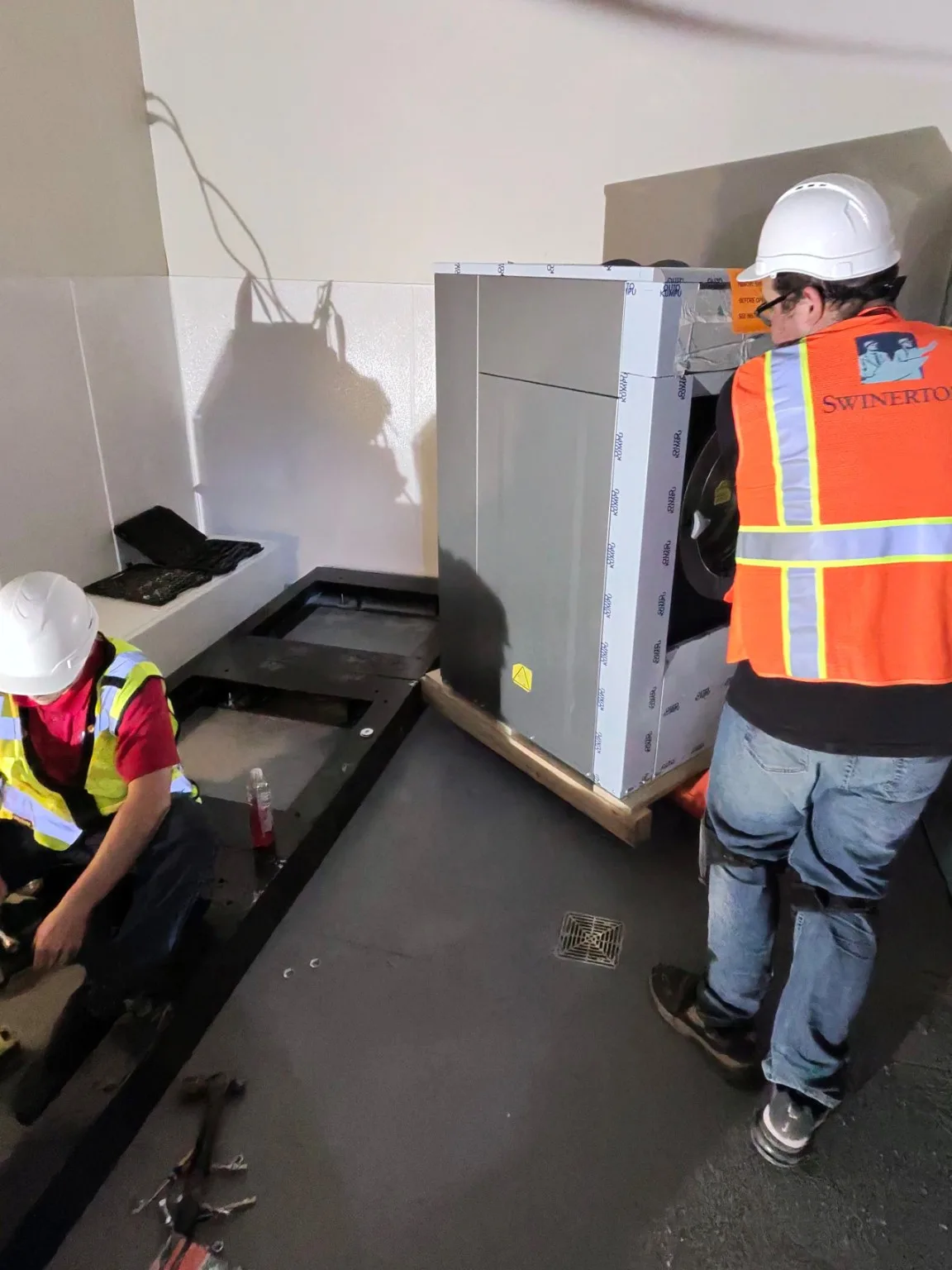 Two technicians in hard hats installing a wrapped commercial washer-extractor on its steel base frame